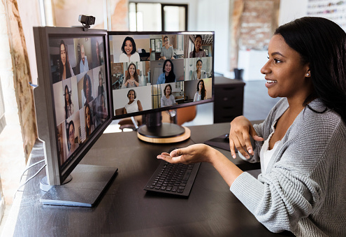 Das Bild zeigt eine junge Frau, die mit ihren Kolleg:innen eine Videokonferenz online an ihrem Computer durchführt. Sie sitzt vor Ihren Bildschirmen, um das pandemiebedingte Social Distancing auch auf Berufswegen einzuhalten.