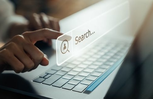 Das Bild zeigt einen Laptop schräg von der Seite. Mithilfe eines gespiegelten Eingabefeldes, erscheint das Wort "Search". Ein Finger deutet darauf zu klicken.