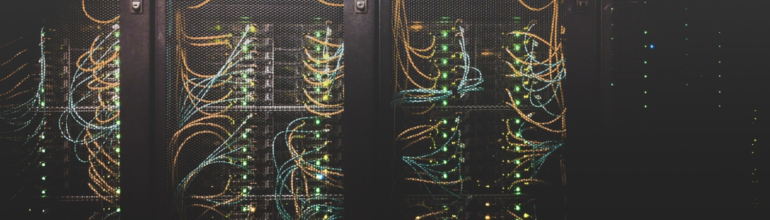Das Bild zeigt die Rückwand eines Servers in transparenten dunklen Tönen, der anhand der bunten Kabel und einigen Leuchteinheiten sichtbar wird.
