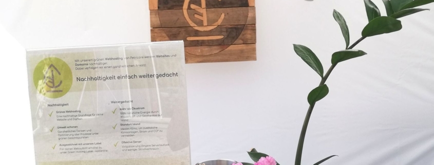 Auf dem Bild ist unser Stand bei der Messe "Green World Tour" von Autarkia in Berlin abgebildet: Darauf ist unser Holzlogo, sowie ein Aufsteller mit unserem Flyer zusehen. Umrandet werden diese von einer Grün- und Trockenpflanze.