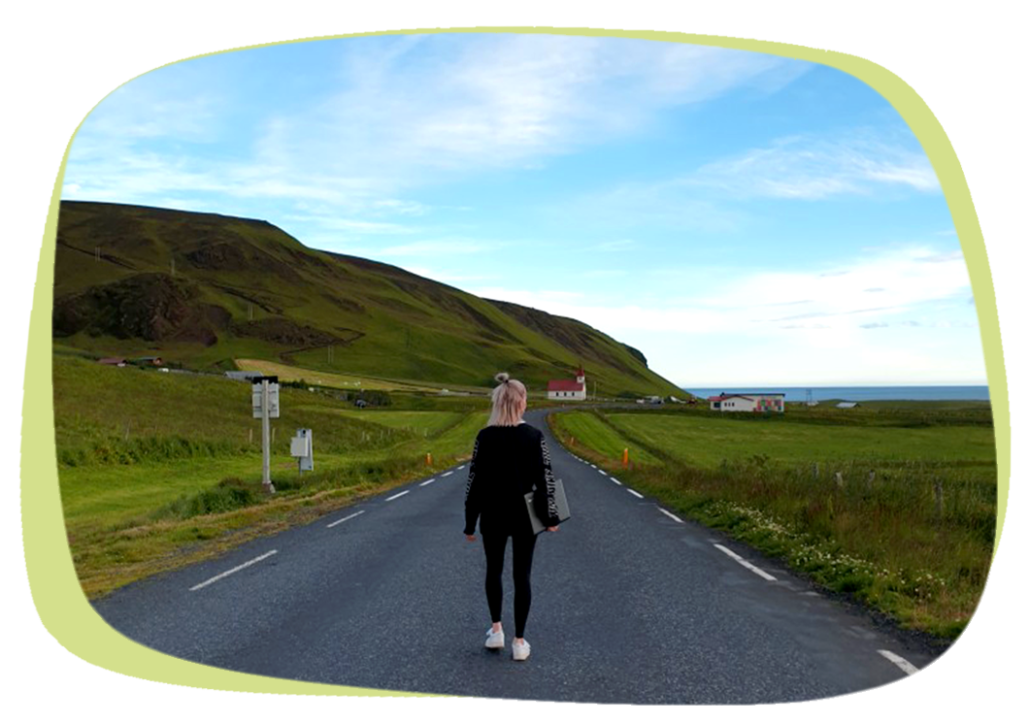 Das Bild zeigt ein Mädchen, das mit dem Laptop unter dem Arm eine Straße in Island entlang läuft. Die Wiesen neben der weiß gestrichelten Straßenbegrenzung zeigen satte Grüntöne. Der Himmel strahlt blau, mit einigen weißen Wolken.