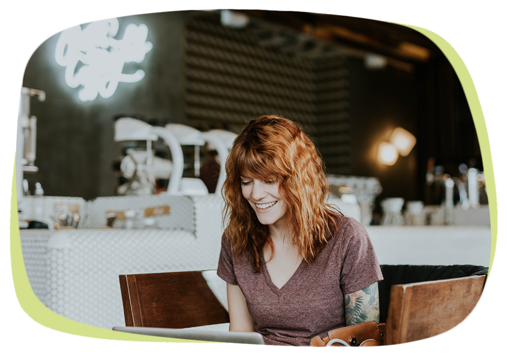 Das Bild rückt eine rothaarige Frau mit einem großen Lächeln in den Fokus. Sie sitzt vor einem aufgeklappten Laptop. Der Hintergrund ist unscharf lässt aber ein Co-Working-Space oder Café vermuten. Das Bild sowie die Freude der jungen, dynamischen Frau steht sinnbildlich für den Service-Gedanken bei Petricore.
