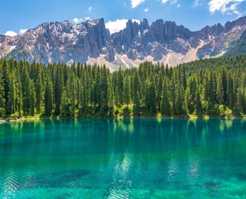 Auf dem Bild ist ein See in Italien mit glasklarem grünblauem und ruhigem Wasser zu sehen. Im Hintergrund sieht man am Ufer einen Wald, dahinter einen bewaldeten Berg und Wolken. Dieses Bild steht sinnbildlich für unseren Blogbeitrag zum Thema Wasser in Europa.