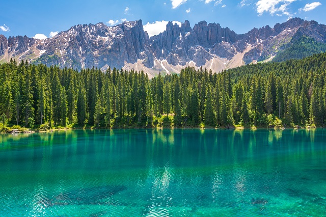 Auf dem Bild ist ein See in Italien mit glasklarem grünblauem und ruhigem Wasser zu sehen. Im Hintergrund sieht man am Ufer einen Wald, dahinter einen bewaldeten Berg und Wolken. Dieses Bild steht sinnbildlich für unseren Blogbeitrag zum Thema Wasser in Europa.