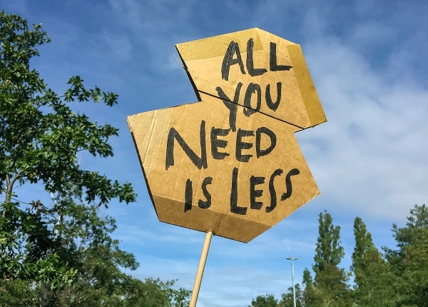 Das Bild zeigt ein Pappschild mit der Aufschrift "All you need is less" in großen Lettern. IM Hintergrund ist blauer Himmel zu sehen und einige Baumwipfel. Der Spruch heißt übersetzt "Alles, was du brauchst, ist weniger." Es steht bei uns sinnbildlich für Emissionen - Denn davon brauchen wir, die Umwelt und die Welt weniger.
