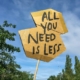 Das Bild zeigt ein Pappschild mit der Aufschrift "All you need is less" in großen Lettern. IM Hintergrund ist blauer Himmel zu sehen und einige Baumwipfel. Der Spruch heißt übersetzt "Alles, was du brauchst, ist weniger." Es steht bei uns sinnbildlich für Emissionen - Denn davon brauchen wir, die Umwelt und die Welt weniger.